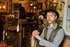 Ein Mann in Steampunk- Optik sitzt in seinem warm und heimelig erleuchteten Museumsraum. Hinter ihm steht ein Tisch mit gläsernen Gefäßen und es hängen Dinge aus vergangenen Zeiten an den Wänden.